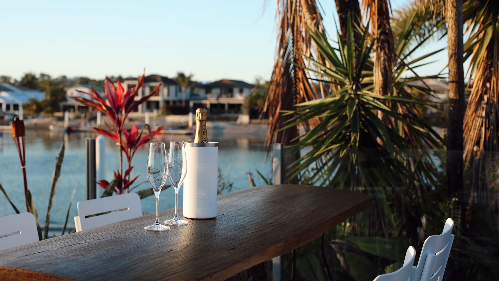 A wooden table with a bottle of champagne and two glasses, overlooking a body of water with houses in the background.