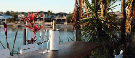A wooden table with a bottle of champagne and two glasses, overlooking a body of water with houses in the background.
