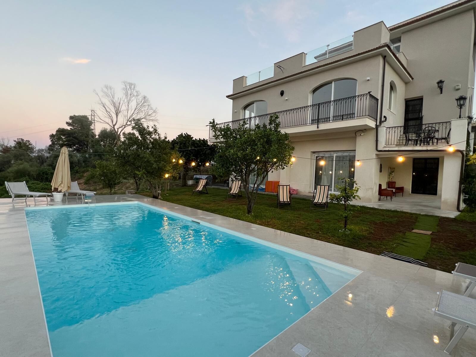 Une maison moderne à deux étages dotée d’une piscine, d’un espace salon extérieur et de guirlandes lumineuses.