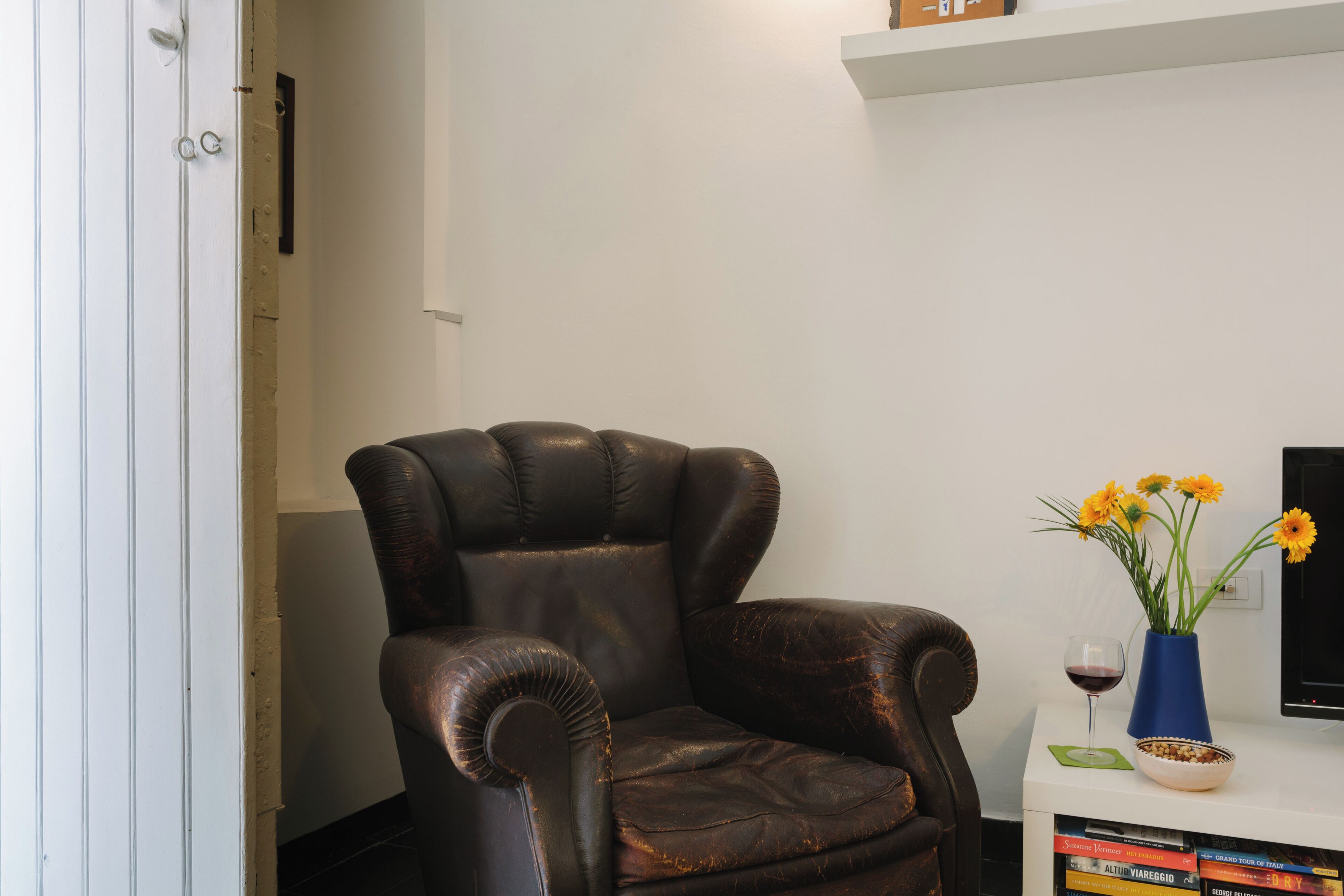 A leather armchair, a vase with flowers, a wine glass, and a stack of books on a white table.