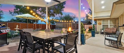 An outdoor patio area with a dining table and chairs, a ceiling fan, string lights, and a fire pit.