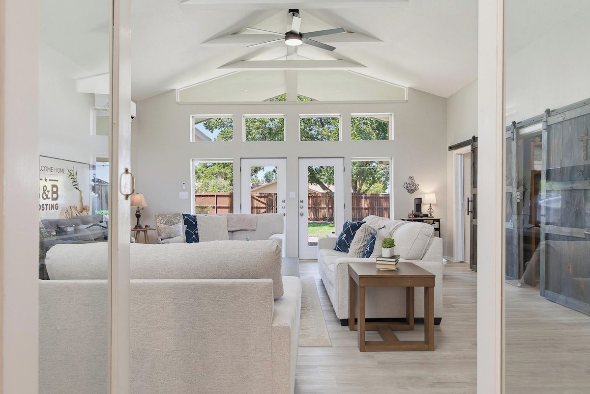 A spacious living room with a high ceiling, a ceiling fan, and a large window overlooking a backyard.