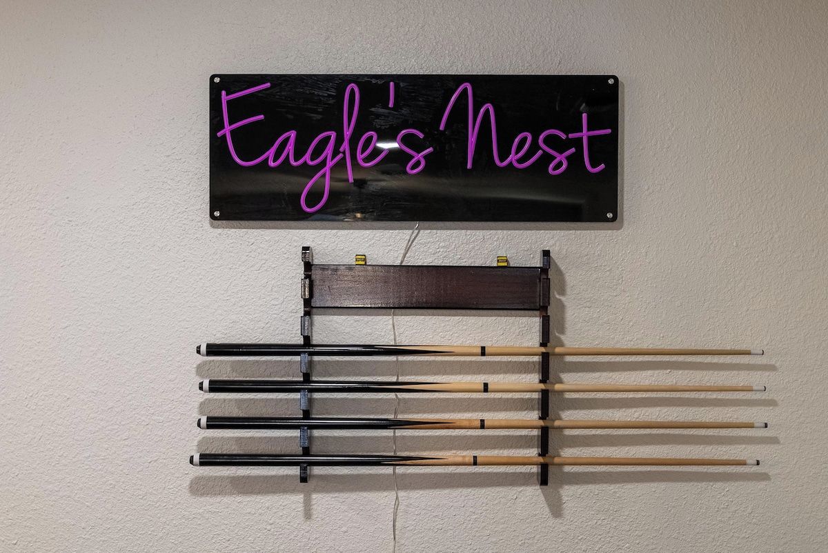 A wall-mounted sign with the text 'Eagle's Nest' and a rack below holding several pool cues.