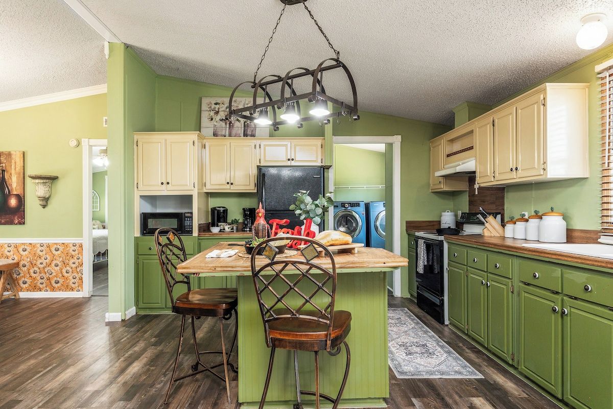 A kitchen with green cabinets, a wooden island with bar stools, a hanging light fixture, and a refrigerator.