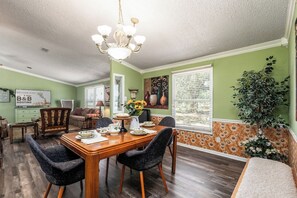 A dining area with a wooden table, chairs, a chandelier, and a large window with blinds.