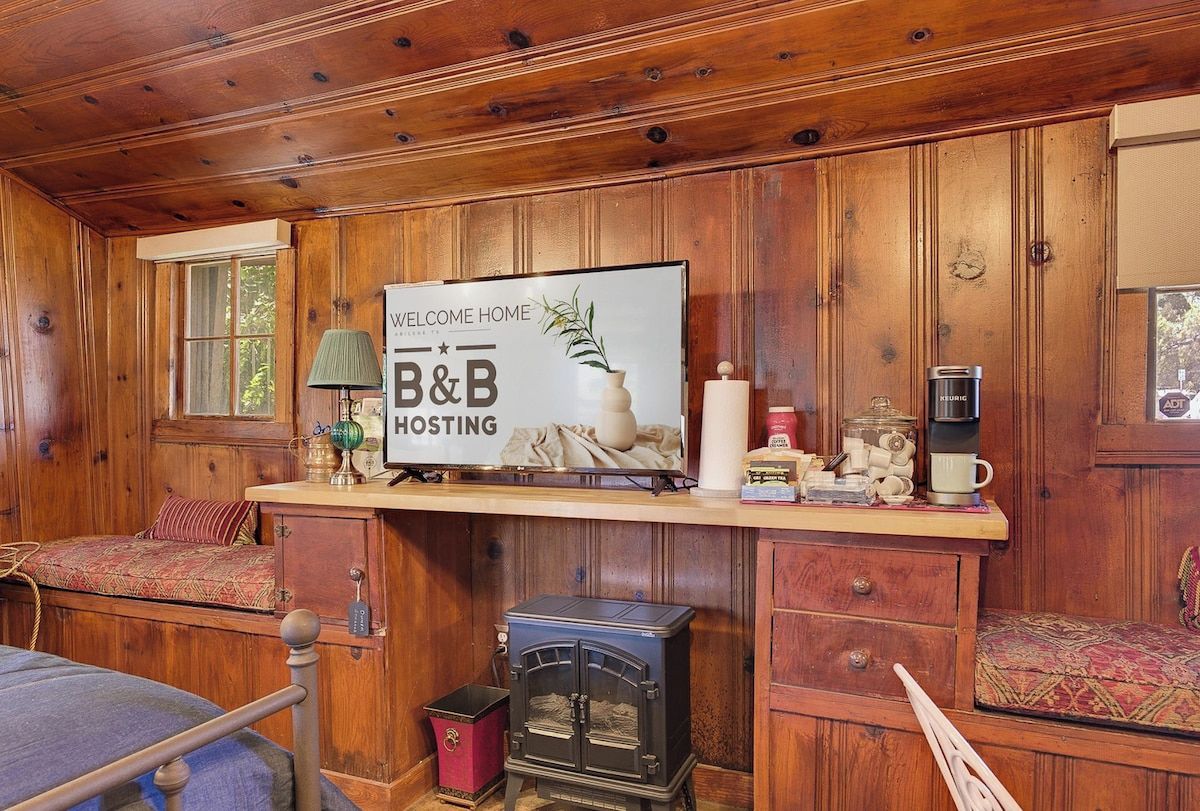 Innenansicht einer Holzhütte mit Bett, einer Couch, einem Kamin, einem Schild mit der Aufschrift 'Welcome Home B&B Hosting' und einer Kaffeemaschine.