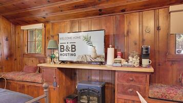 Innenansicht einer Holzhütte mit Bett, einer Couch, einem Kamin, einem Schild mit der Aufschrift 'Welcome Home B&B Hosting' und einer Kaffeemaschine.