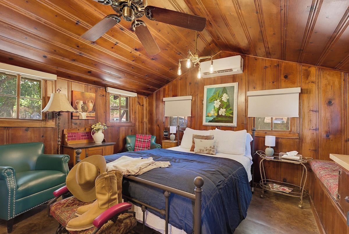 A wooden-paneled room with a bed, a green armchair, a small table, and a ceiling fan.