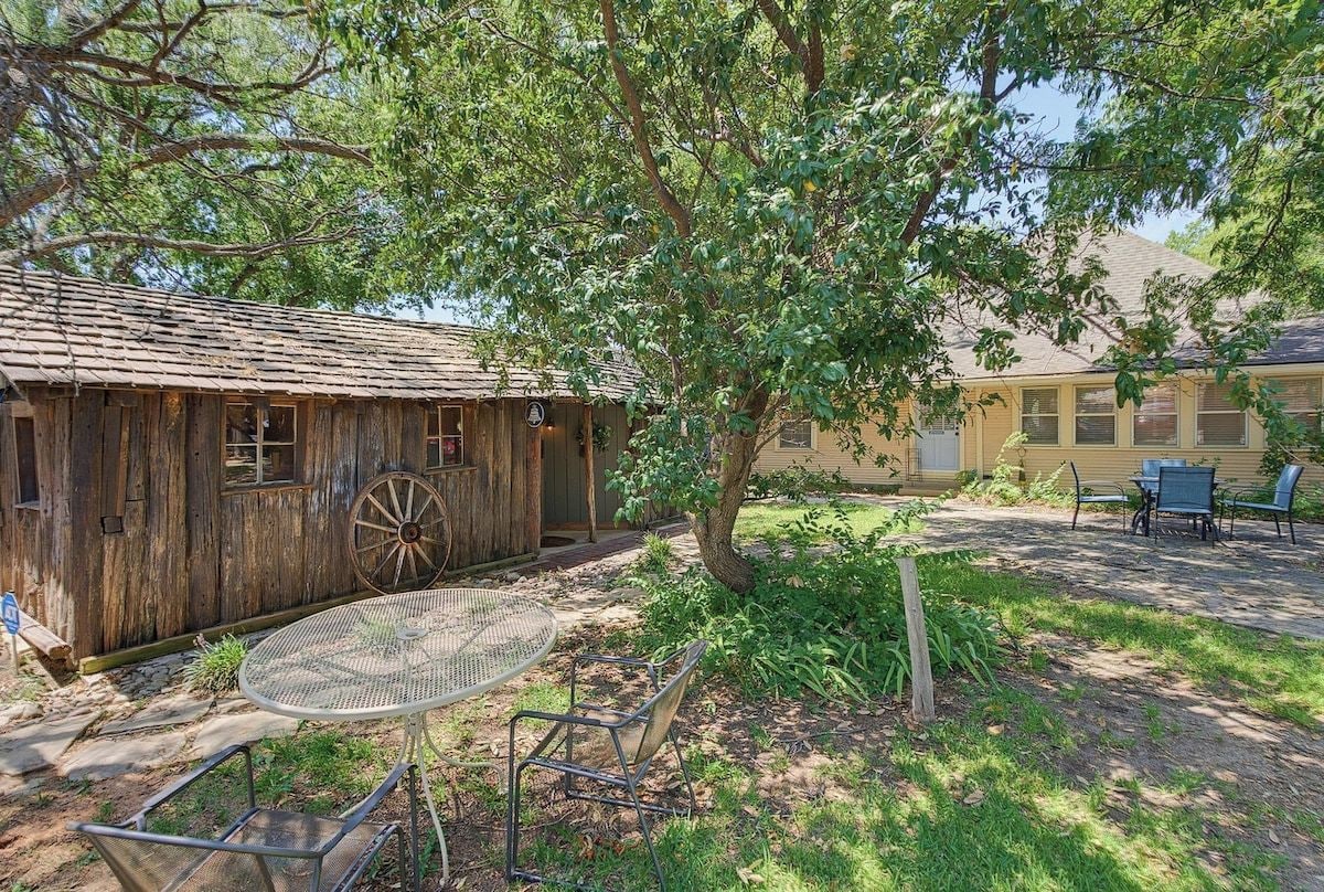 A rustic wooden building with a large wheel, a metal table with chairs, and a garden area.