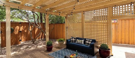 Un patio exterior con una pérgola de madera, un sofá azul, una mesa de comedor con sillas y plantas en macetas.
