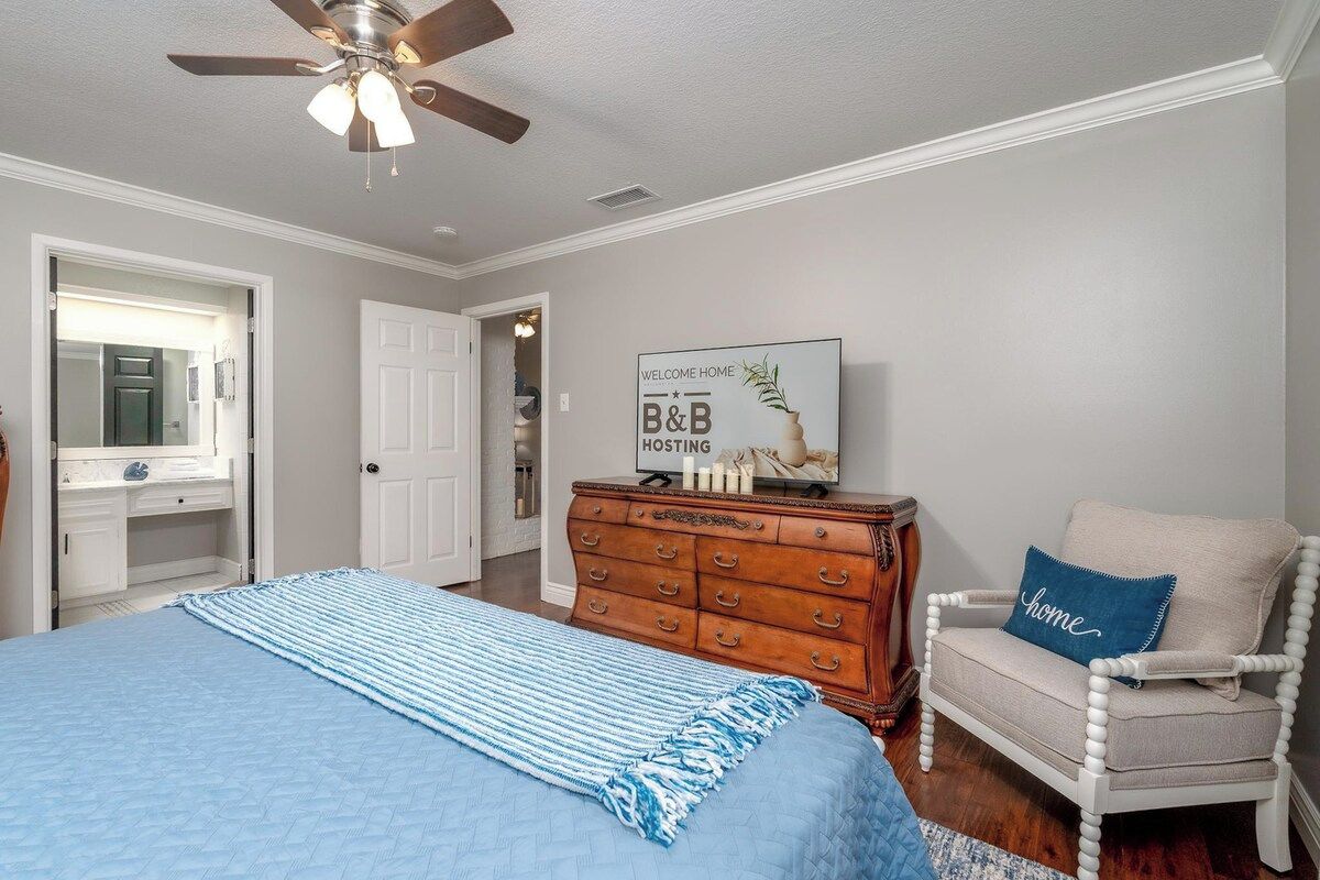 A bedroom with a bed, a dresser, a chair, a ceiling fan, and a welcome home sign.