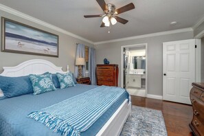 Ein Schlafzimmer mit Bett, Deckenventilator, Kommode, Nachttisch, Lampe, einem Gemälde und einer Tür zu einem Badezimmer.