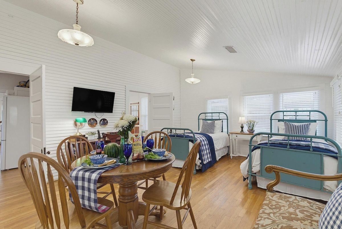 A room with a dining table, chairs, a bed with a metal frame, a television mounted on the wall, and a window with blinds.