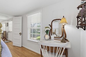 A room with a white door, a window with white blinds, a white vanity with a mirror, a brass lamp, and a wooden chair.