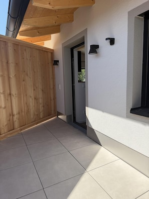 A covered outdoor area with a wooden fence, a tiled floor, and a white wall with a window and wall-mounted lights. - Limone Apartment with Rooftop Terrace (Bernau am Chiemsee)