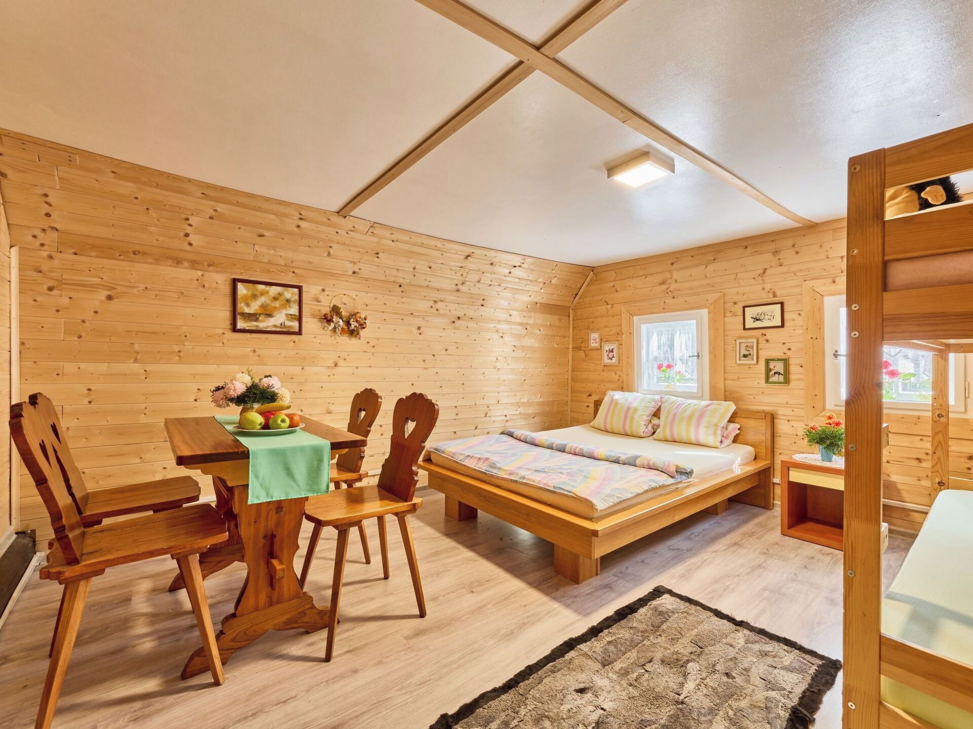 A cozy wooden cabin interior with a bed, a dining table with chairs, and a small table with flowers.