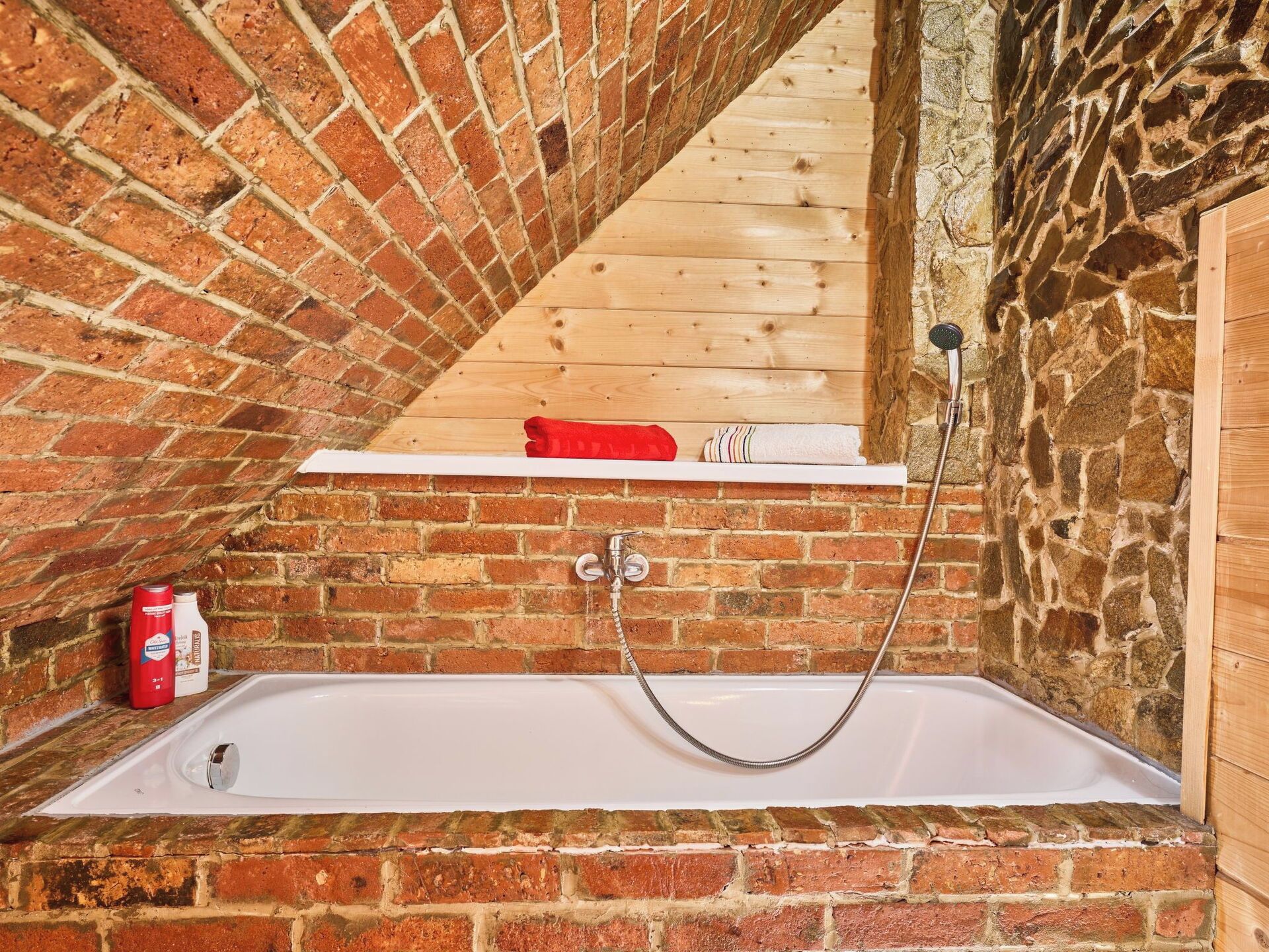 Une salle de bain avec un mur en briques, une étagère en bois, une baignoire et une pomme de douche.