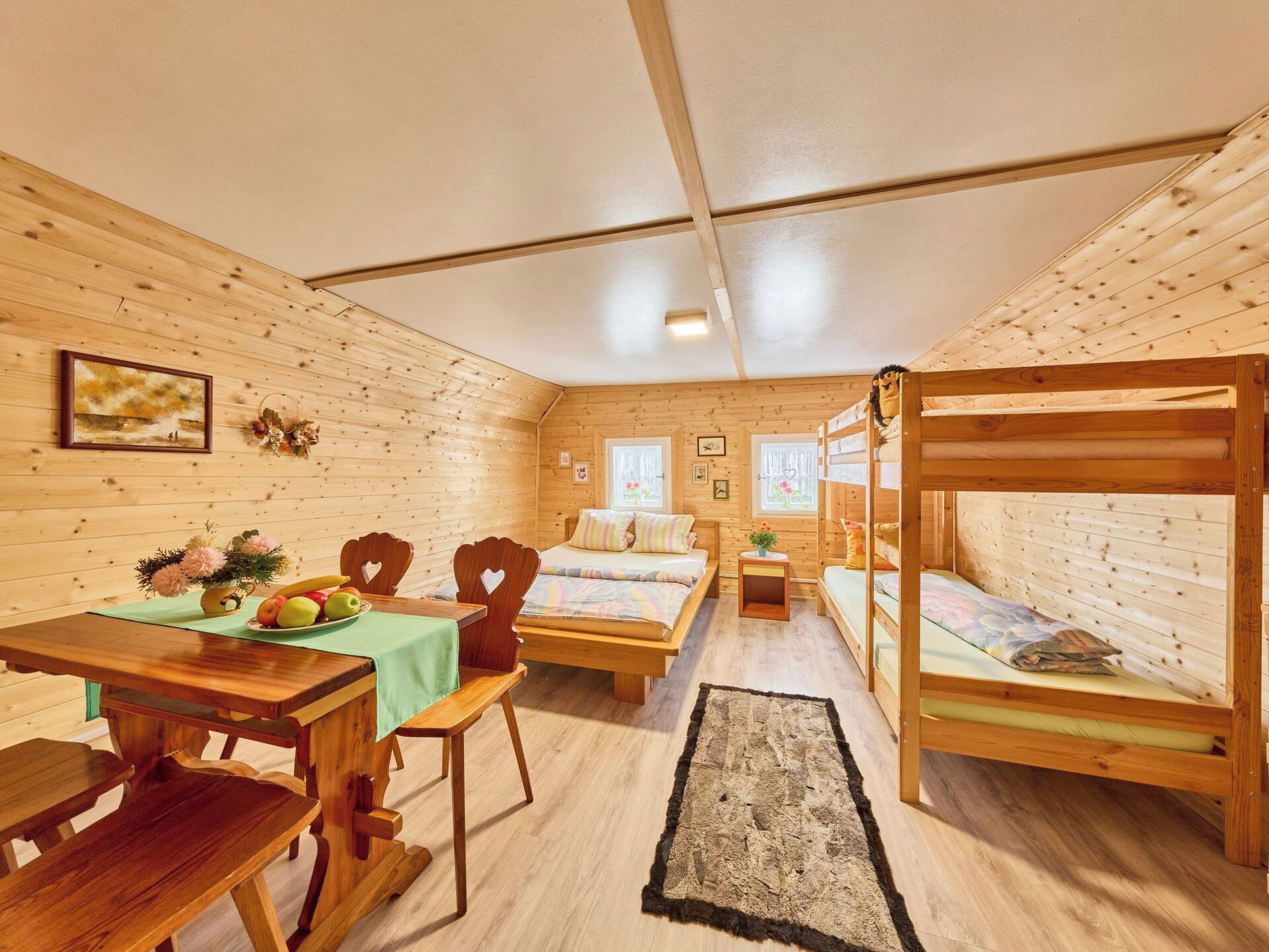 A cabin interior with wooden walls, a dining table with chairs, a bed with a headboard, and a bunk bed.