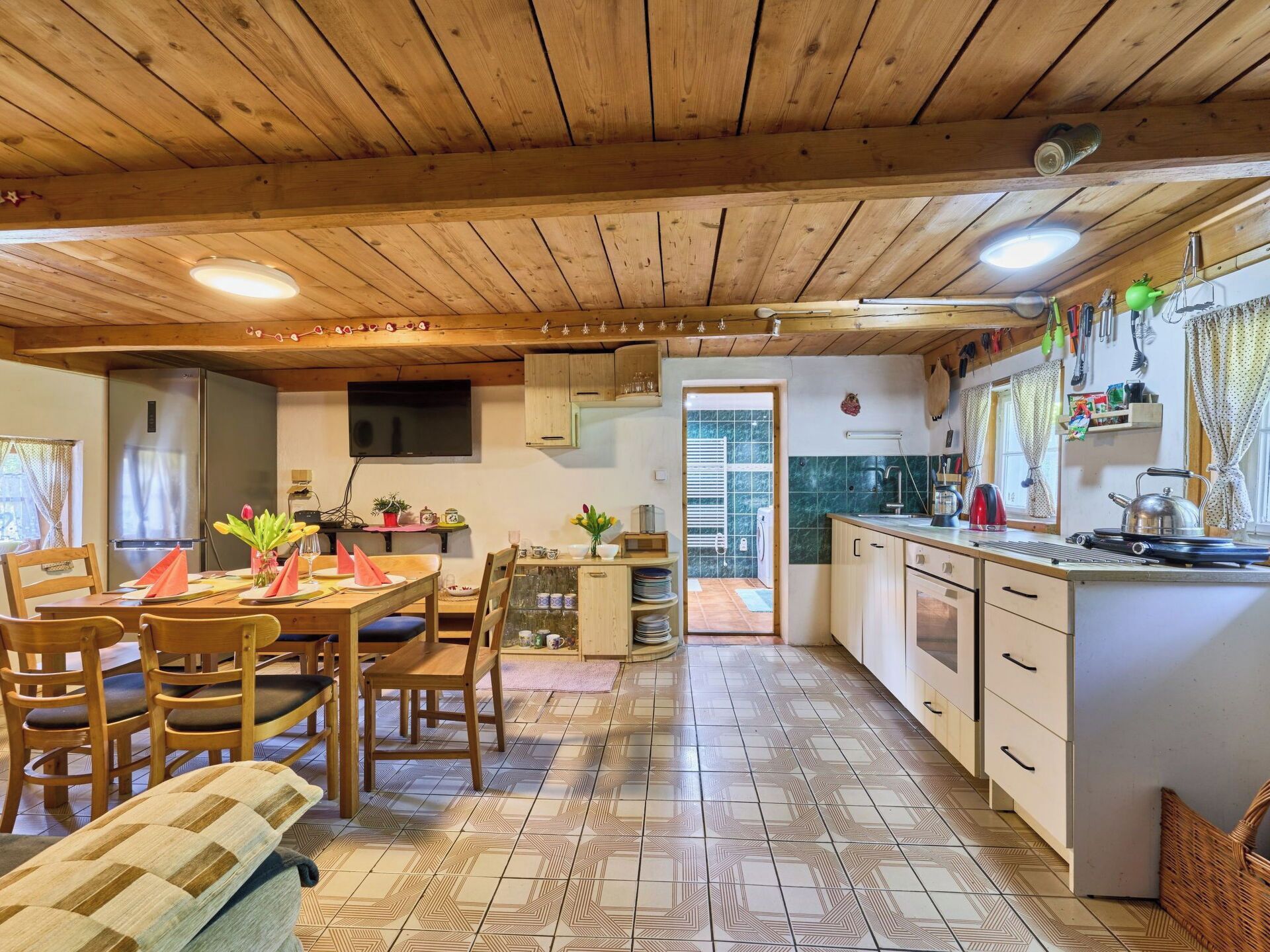 A kitchen with wooden cabinets, a dining table with chairs, a refrigerator, and a television mounted on the wall.
