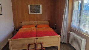 A bedroom with a wooden bed, a nightstand, a chair, a radiator, and a window with curtains. - Birke, (Grächen), by Interhome (Grächen)