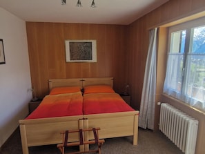 A bedroom with a wooden bed, a nightstand, a chair, a radiator, and a window with curtains. - Birke, (Grächen), by Interhome (Grächen)