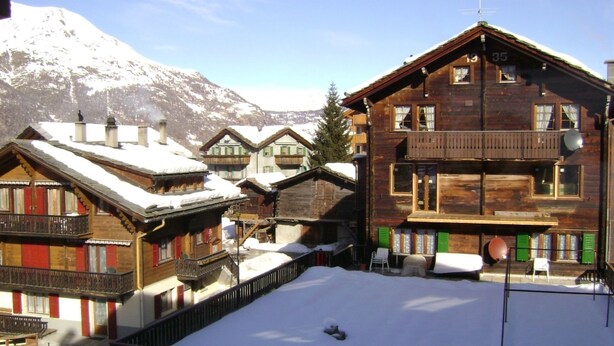 Een houten chalet met balkon, omringd door besneeuwde gebouwen, met een berg op de achtergrond.