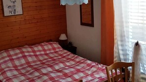 Een kamer met een houten wand, een bed met een geruit beddegoed, een hanglamp, een ingelijst schilderij en een raam met gordijnen.