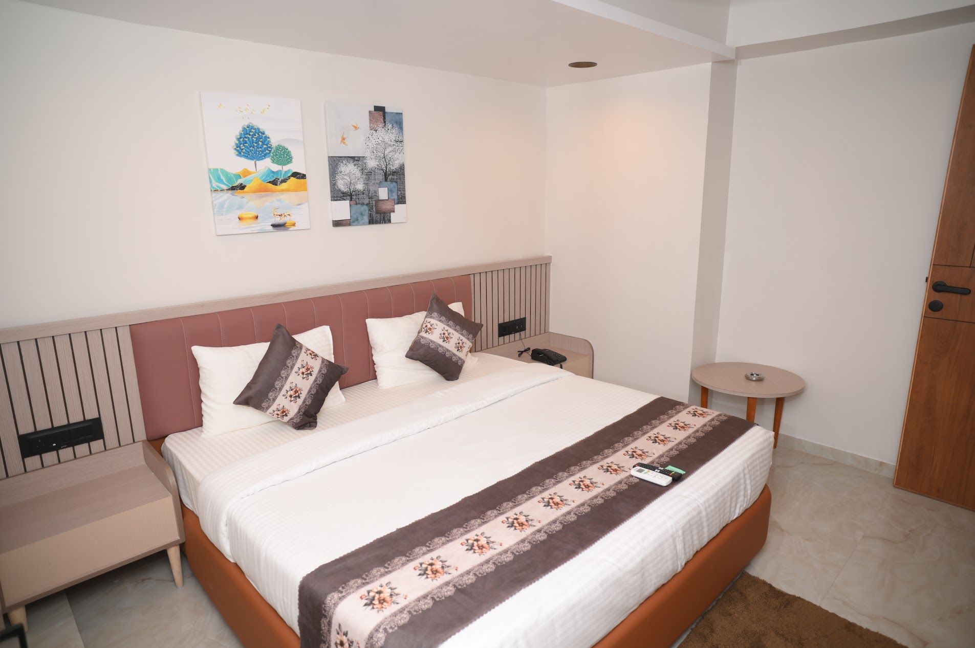 A neatly arranged hotel room with a bed, bedside tables, a small round table, and two wall-mounted artworks.