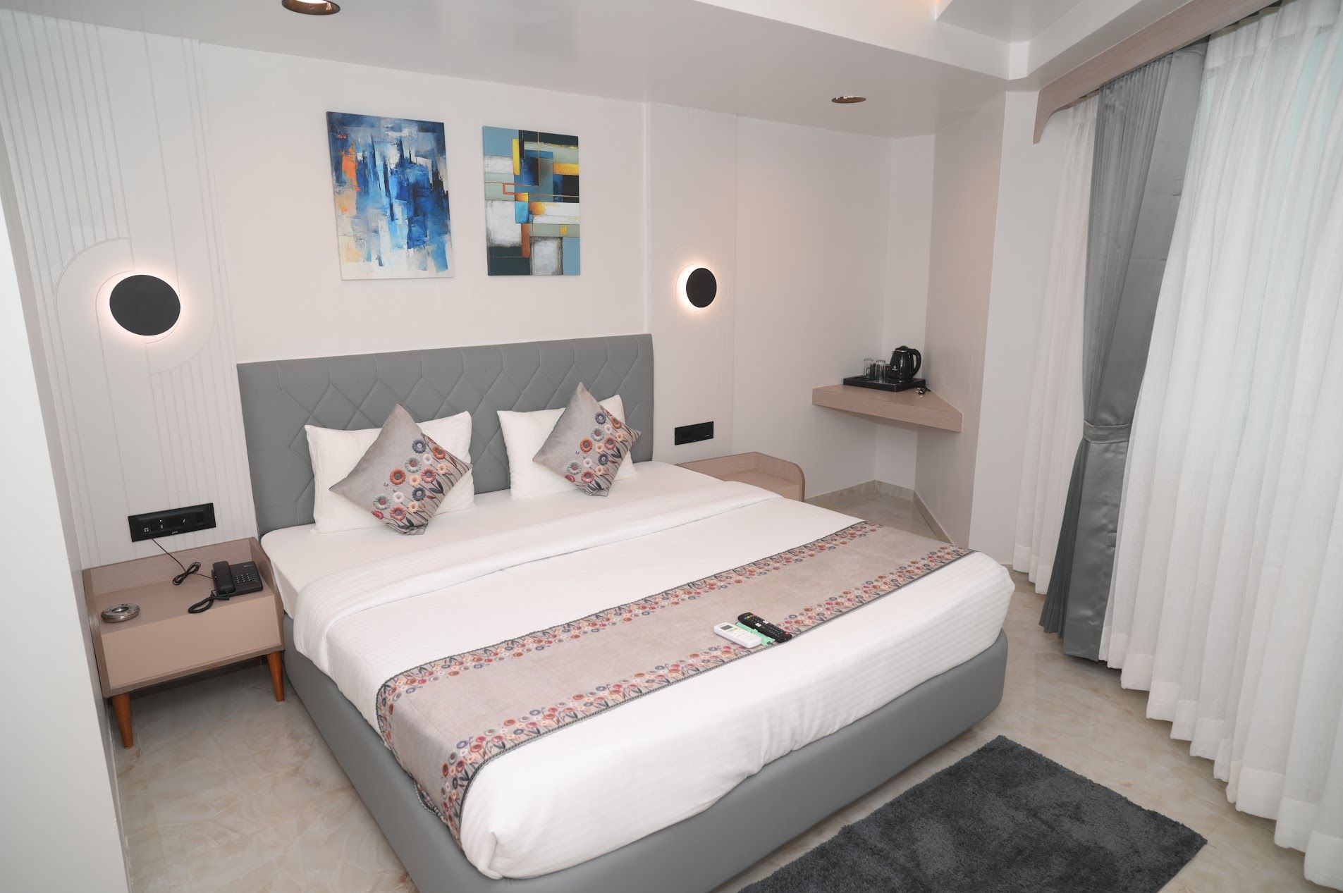 A modern hotel room with a large bed, two bedside tables, a bedside lamp, a nightstand, a wall-mounted phone, a small shelf with a kettle, and a window with curtains.