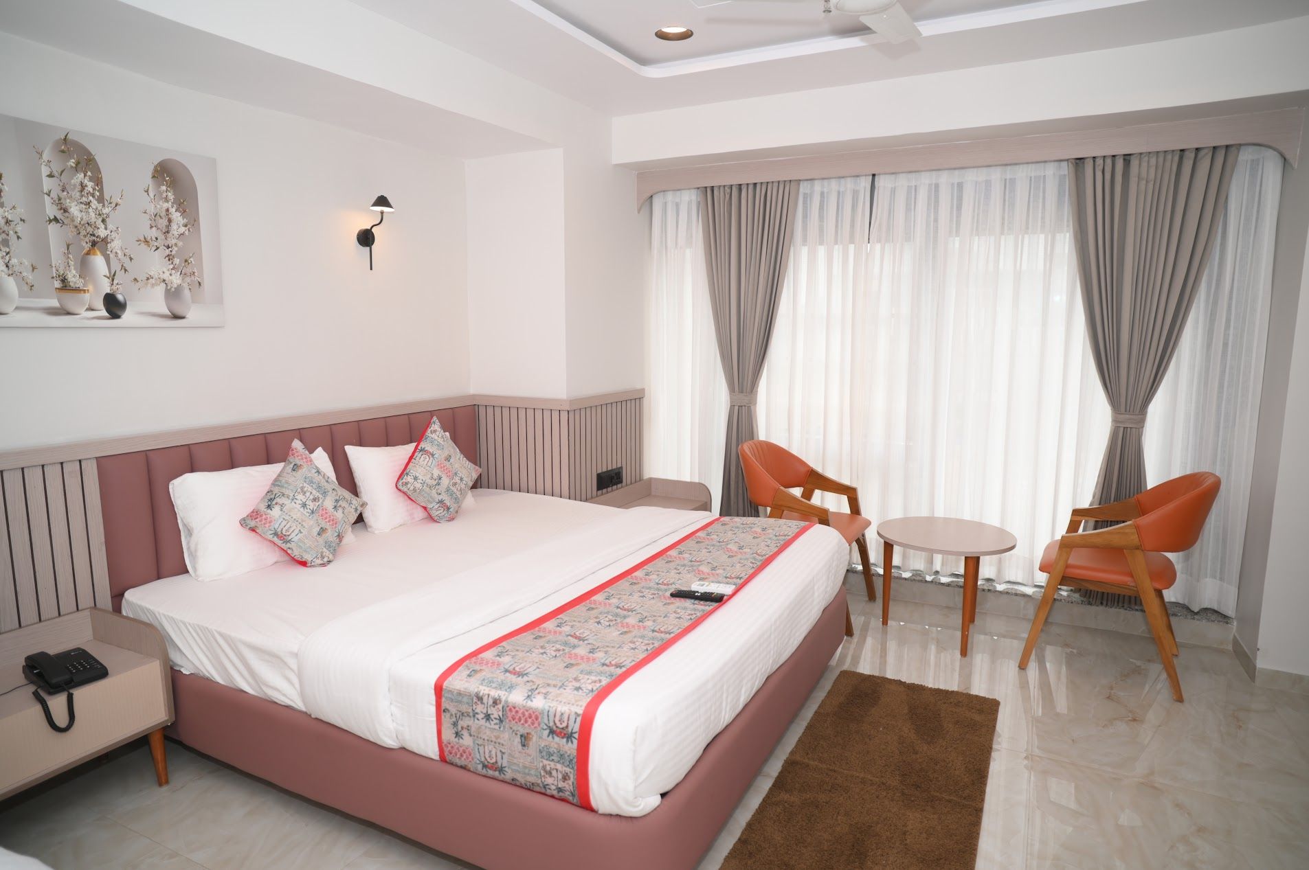 A hotel room with a large bed, a bedside table with a phone, two chairs, a small table, and a large window with curtains.