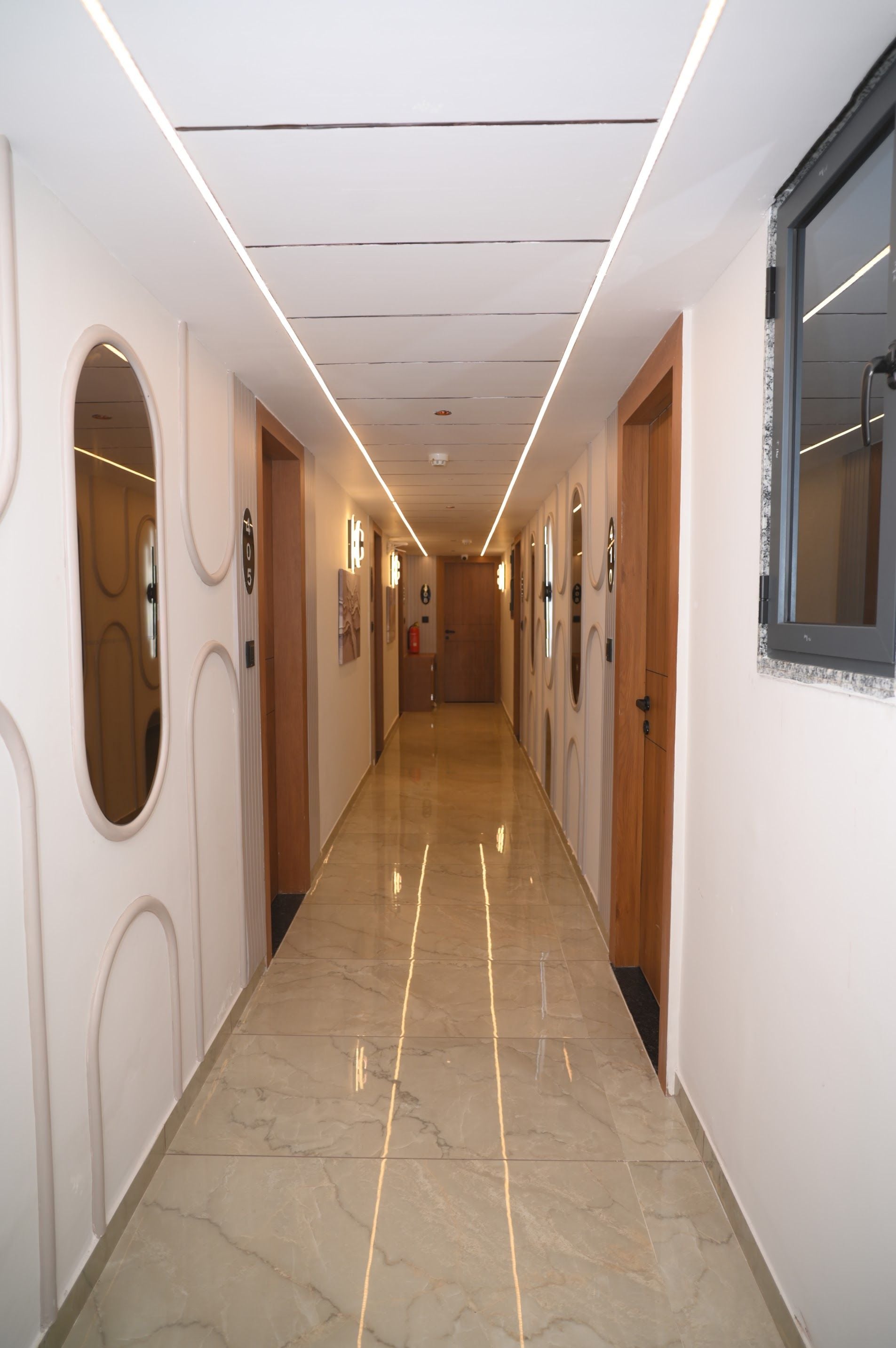Un couloir d’hôtel avec un sol en marbre, des portes en bois et des luminaires encastrés au plafond.