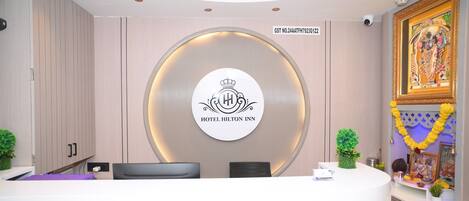 Un mostrador de recepción de hotel con un logo circular, una cámara de seguridad y una pared decorativa con un cuadro enmarcado.