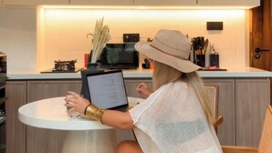 Una persona sentada frente a una mesa redonda con una computadora portátil, en una habitación con armarios de madera y una cocina al fondo.