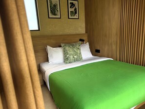Ein ordentlich bezogenes Bett mit einer grünen Tagesdecke und zwei Kissen, eines mit Blätermotiv, in einem Zimmer mit Holzvertäfelung und gerahmten Pflanzenbildern an der Wand.
