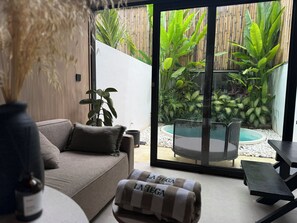 A modern living room with a sofa, a bench, and a small table. There is a large sliding glass door leading to an outdoor area with plants and a seating area. - Private Jungle Pool Loft Near Canggu Beach • Guest Favorite • Fast WiFi (Canggu)