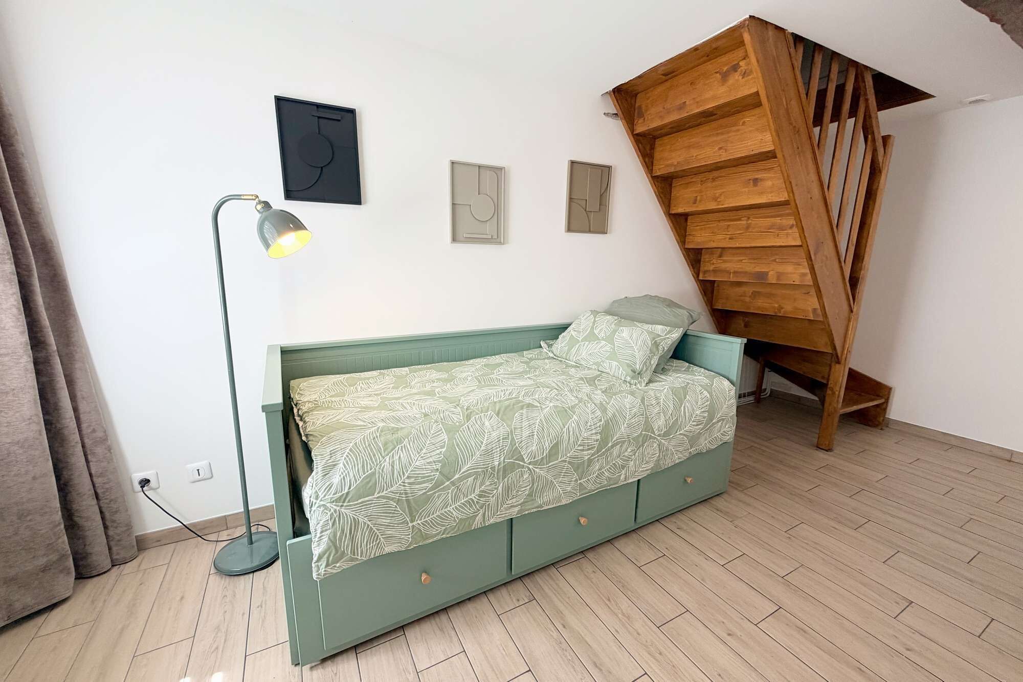 A room with a bed, a standing lamp, a wall-mounted artwork, and a wooden staircase.