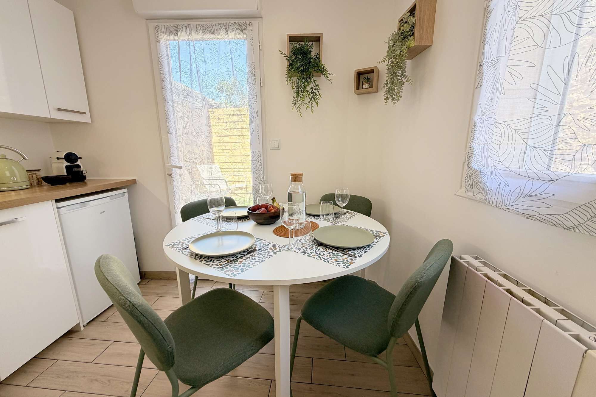 A dining area with a round table, chairs, and a view of the outdoors through a window with a leaf-patterned curtain.
