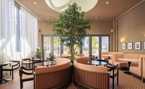 A modern indoor seating area with round tables and cushioned seating, a large tree, and large windows. - The Crosby Balcony Studio Miami Worldcenter (Miami)