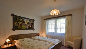 Ein Schlafzimmer mit einem Doppelbett, zwei Nachttischlampen, einem Nachttisch, einem Sessel, einem Fenster mit Vorhängen und einem Wandbild.