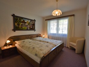 Ein Schlafzimmer mit einem Doppelbett, zwei Nachttischlampen, einem Nachttisch, einem Sessel, einem Fenster mit Vorhängen und einem Wandbild.