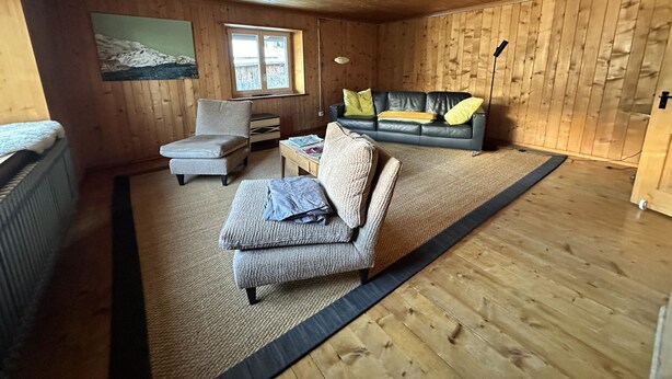A cozy cabin interior with a wooden ceiling, a sofa, a chair, a small table, and a window.