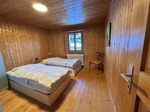 Ett sovrum i en stuga, inrett med träpanel, och det innehåller två sängar, ett element, ett fönster och en stol.