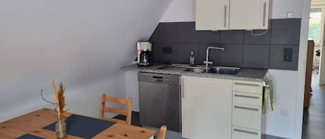 A compact kitchen with a dishwasher, a coffee maker, and a sink. There is a dining table with chairs and a vase of dried flowers.