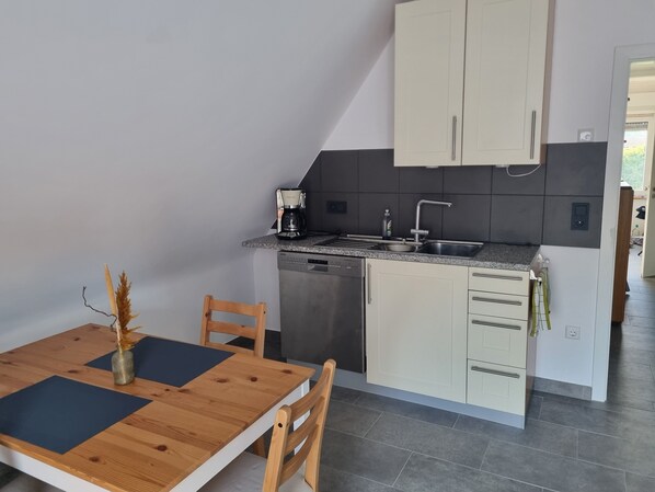 A compact kitchen with a dishwasher, a coffee maker, and a sink. There is a dining table with chairs and a vase of dried flowers.