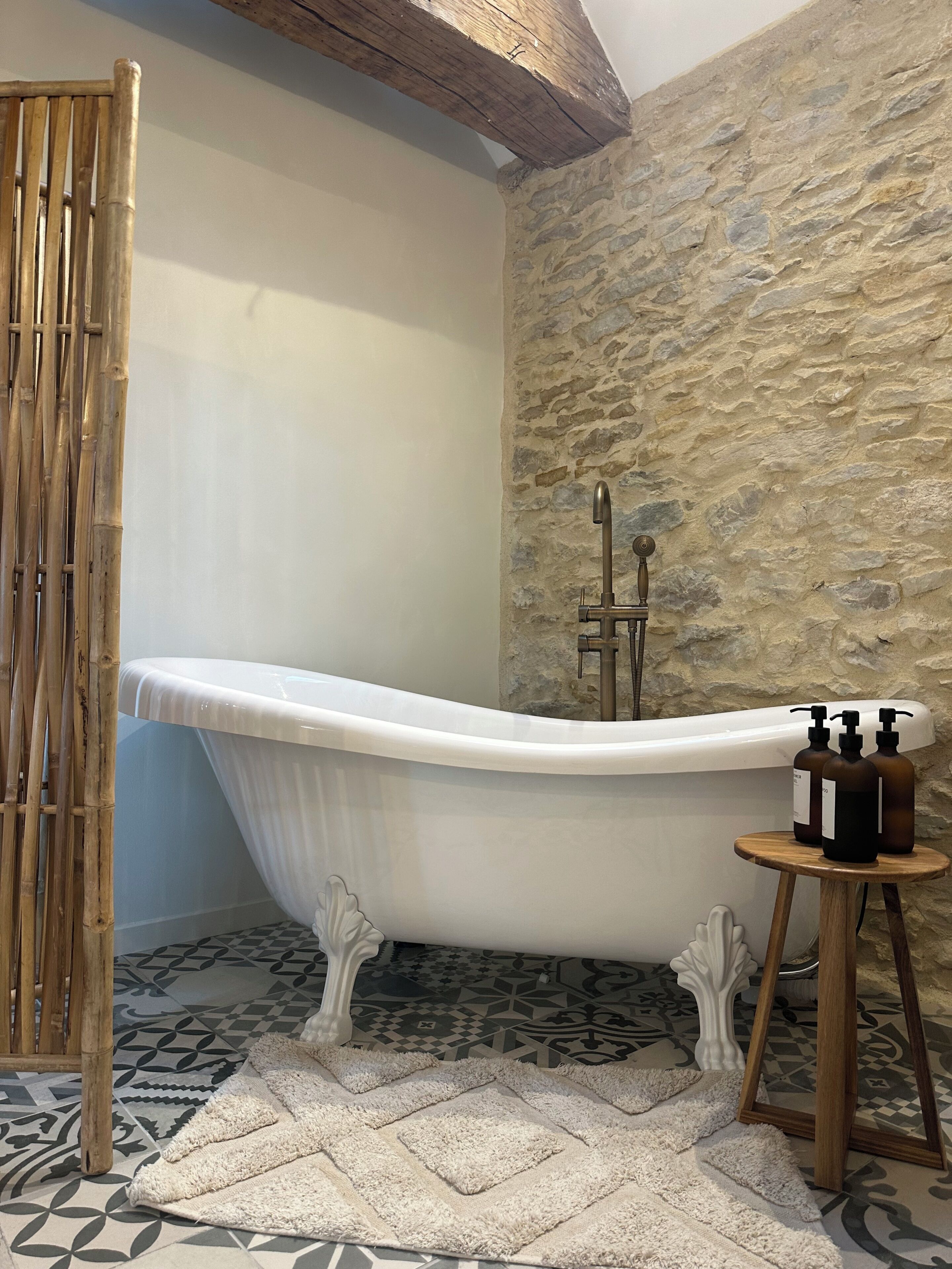 Une salle de bain avec une baignoire sur pieds, un mur en pierre, une décoration en bambou, des carreaux de sol à motifs et un petit tabouret en bois sur lequel sont posées des bouteilles.