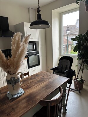 A modern kitchen with white cabinetry, a wooden dining table, and a view of the outside through a window. - Le Clos des Murmures - maisonnette mitoyenne à Trept (Trept)
