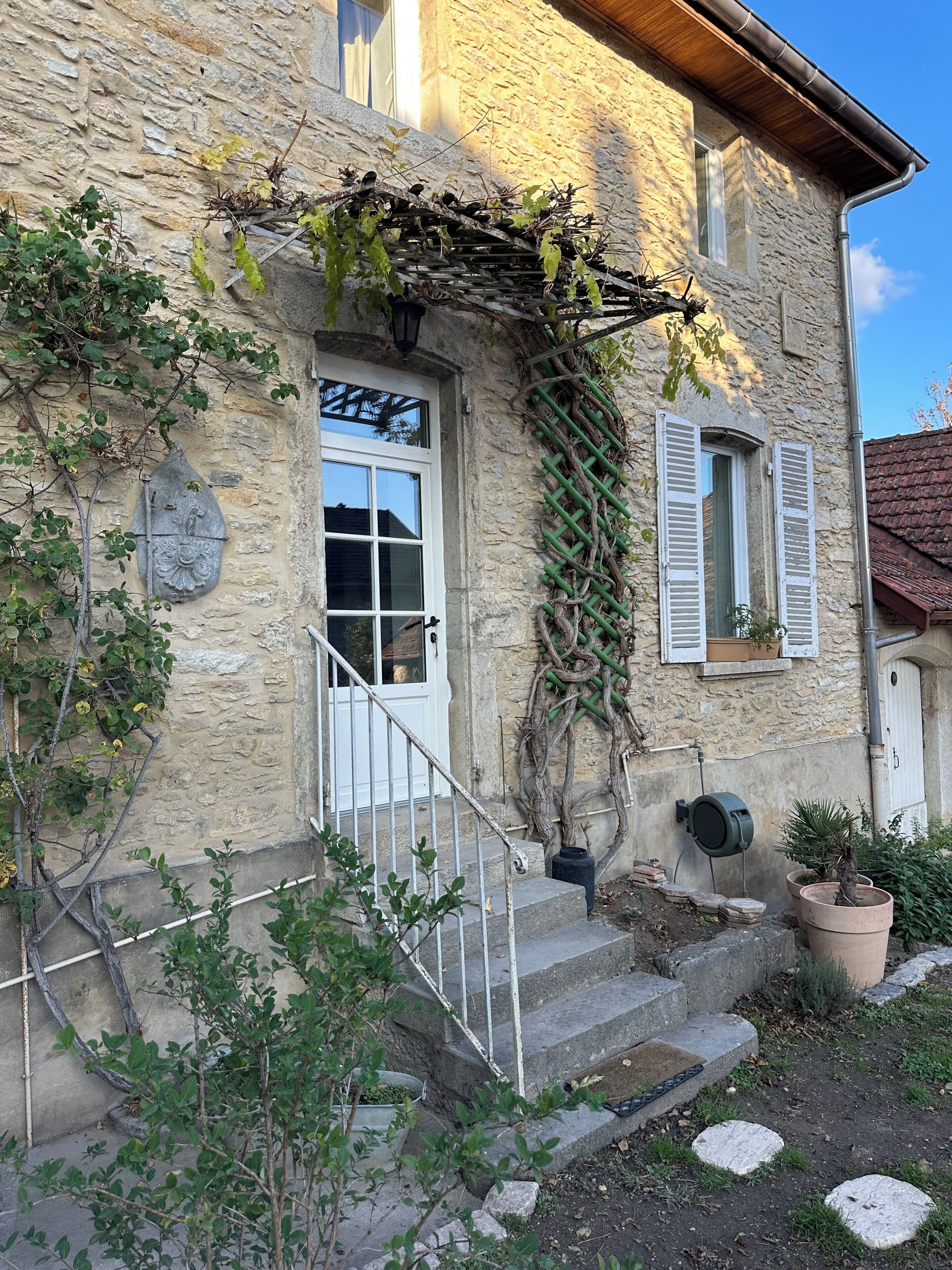 Ein Steinhaus mit einer weißen Tür, einer kleinen Treppe, die zum Eingang führt, und einem Gartenbereich mit Topfpflanzen.