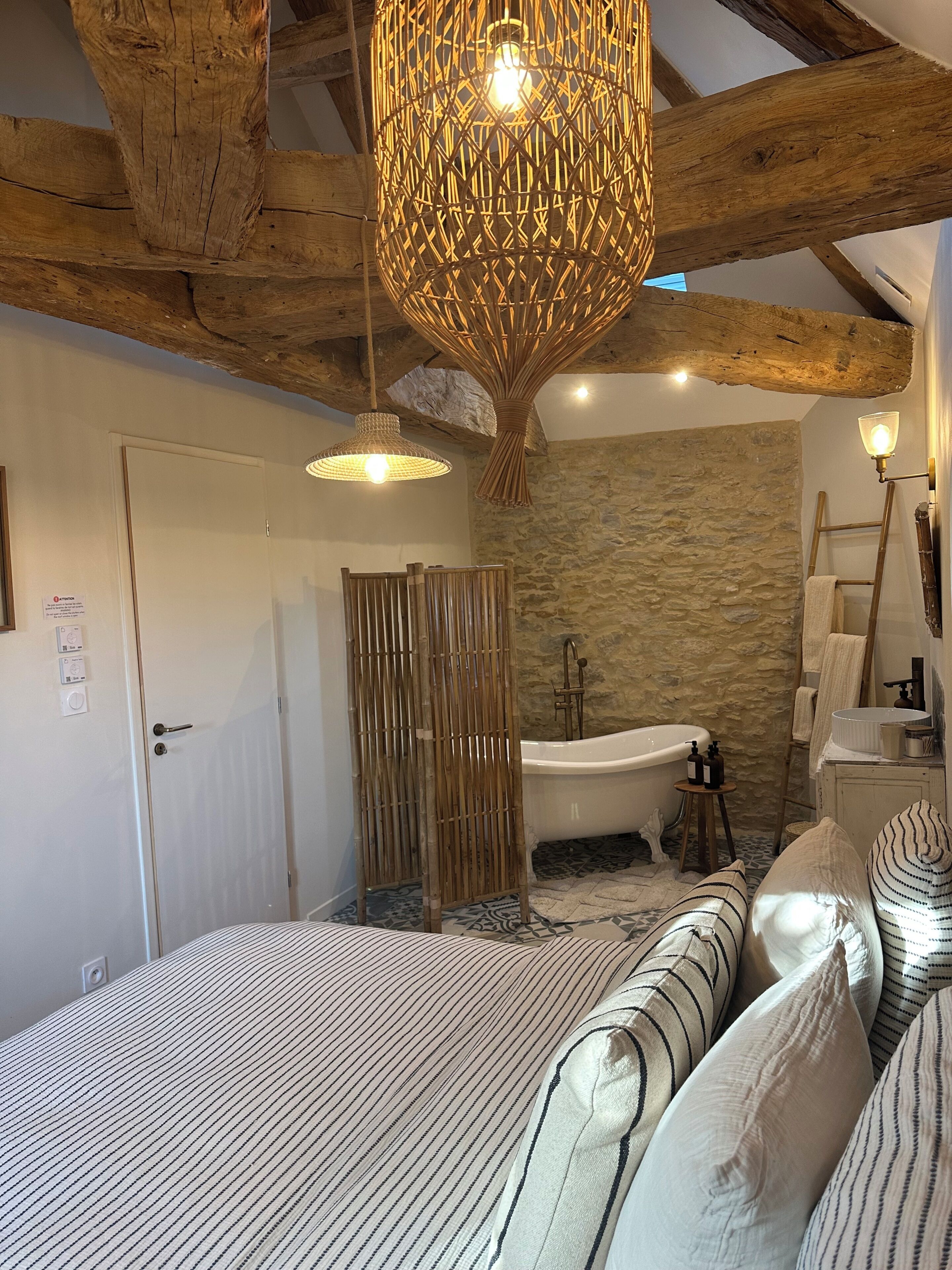 A bedroom with a large bed, a wooden ceiling, a stone wall, a bathtub, and a hanging light fixture.