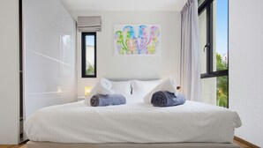 A neatly made bed with two rolled towels, a window with curtains, and a colorful abstract painting on the wall. - Stylish 1BR Apartment at The Title Halo, Nai Yang (Tambon Sa Khu)