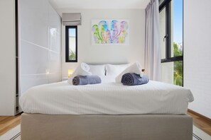 Ein ordentlich bezogenes Bett mit zwei gerollten Handtüchern, ein Fenster mit Vorhängen und ein farbenfrohes abstraktes Bild an der Wand.